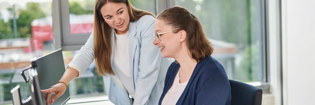 Zwei Frauen im Büro arbeiten gemeinsam am Computer im Bereich digitaler Vertrieb. Die stehende Frau zeigt auf den Bildschirm und erklärt der sitzenden Frau digitale Vertriebsstrategien. Helle, freundliche Büroumgebung mit großen Fenstern im Hintergrund. Das Bild symbolisiert Teamwork, digitale Verkaufsprozesse und den Einsatz moderner Technologien im Vertrieb.