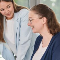 Zwei Frauen im Büro arbeiten gemeinsam am Computer im Bereich digitaler Vertrieb. Die stehende Frau zeigt auf den Bildschirm und erklärt der sitzenden Frau digitale Vertriebsstrategien. Helle, freundliche Büroumgebung mit großen Fenstern im Hintergrund. Das Bild symbolisiert Teamwork, digitale Verkaufsprozesse und den Einsatz moderner Technologien im Vertrieb.